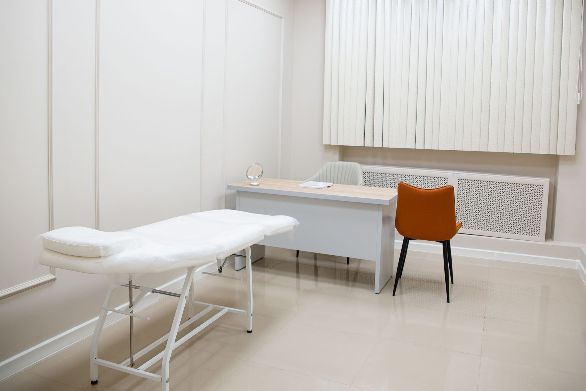 A minimalist medical examination room featuring a desk, chairs, and an examination bed in a sterile, neutral setting.